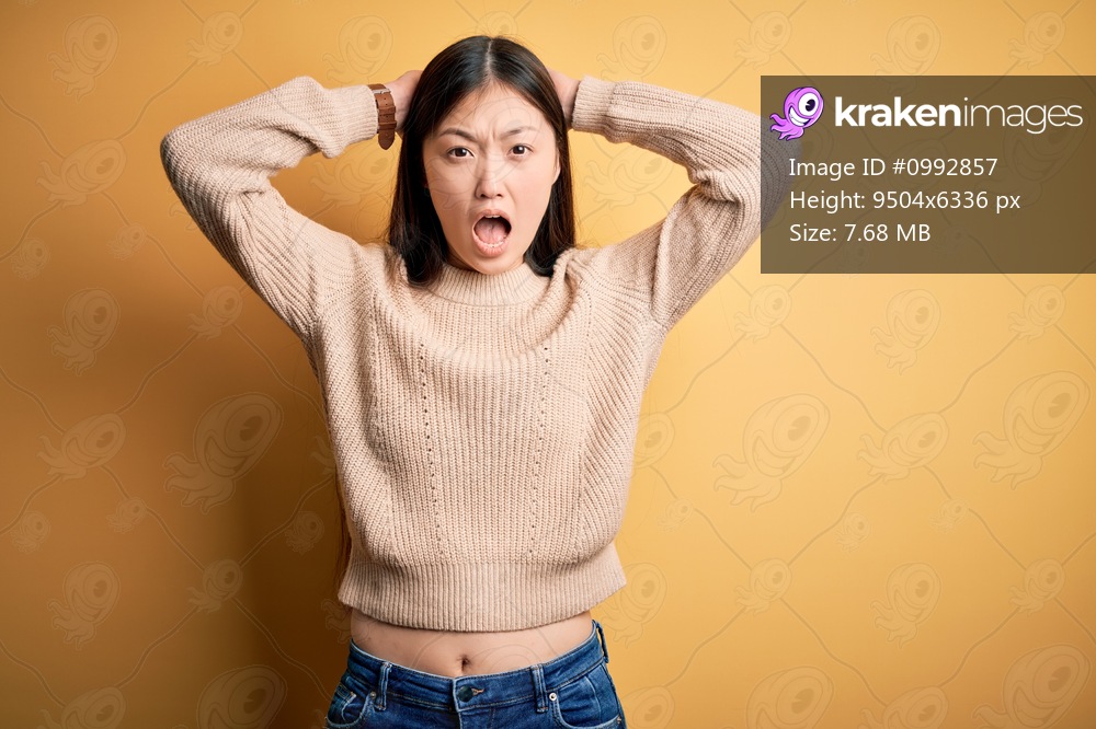 Young beautiful asian woman wearing casual sweater over yellow isolated background Crazy and scared with hands on head, afraid and surprised of shock with open mouth