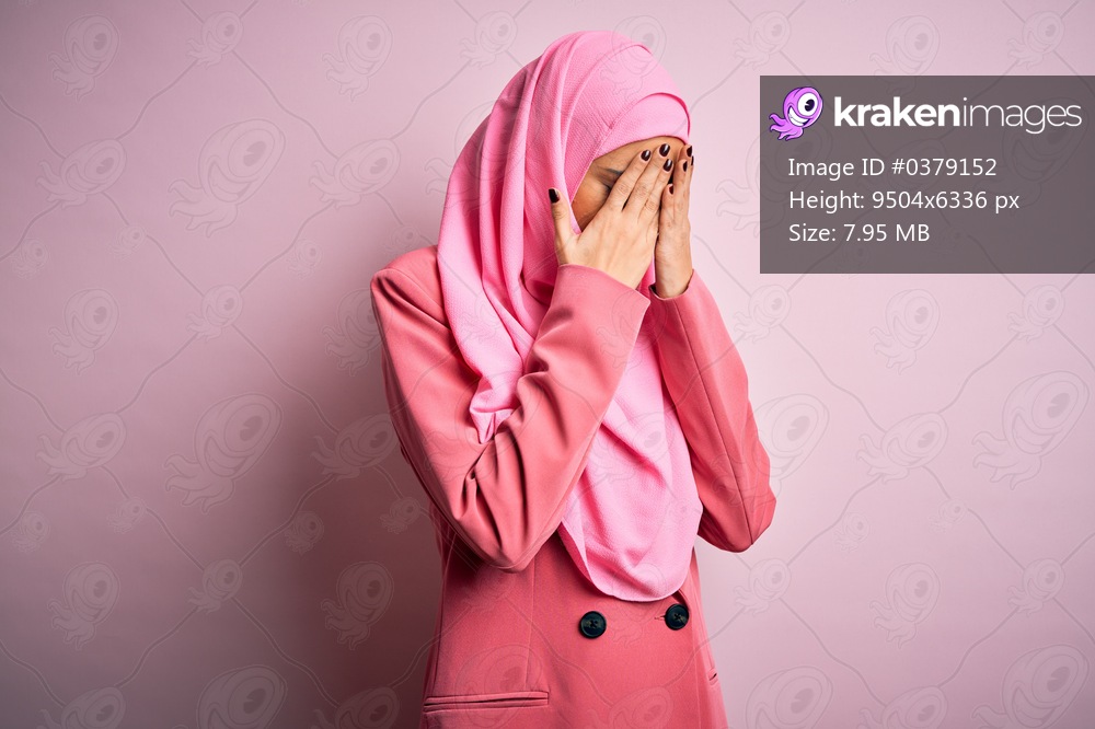 Young beautiful brunette businesswoman wearing pink muslim hijab and business jacket with sad expression covering face with hands while crying. Depression concept.