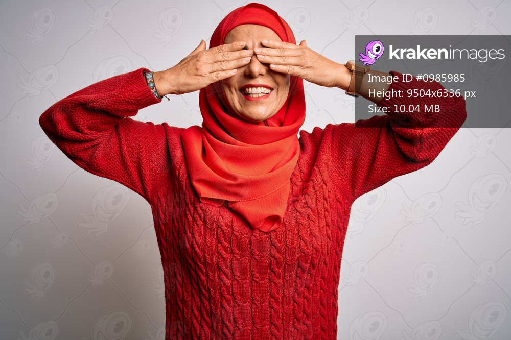 Middle age brunette woman wearing muslim traditional hijab over isolated white background covering eyes with hands smiling cheerful and funny. Blind concept.