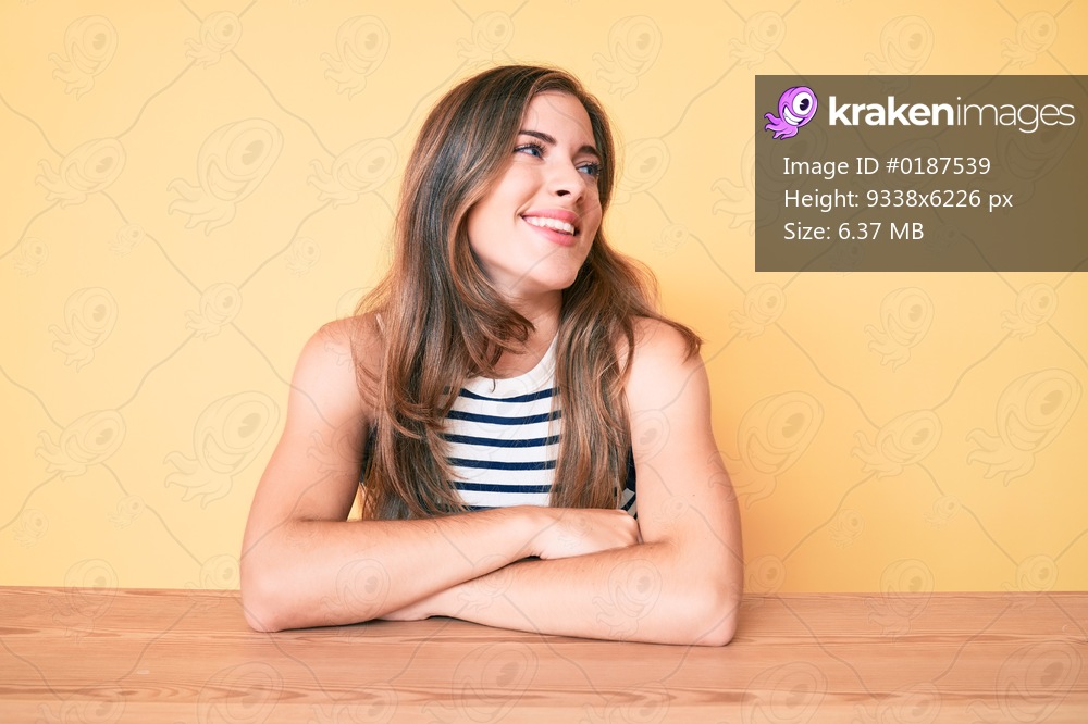 Beautiful young caucasian woman wearing casual clothes sitting on the table looking away to side with smile on face, natural expression. laughing confident. 