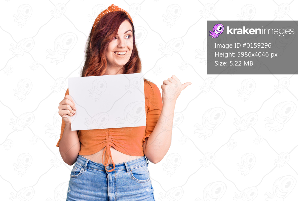 Young beautiful woman holding cardboard banner with blank space pointing thumb up to the side smiling happy with open mouth 