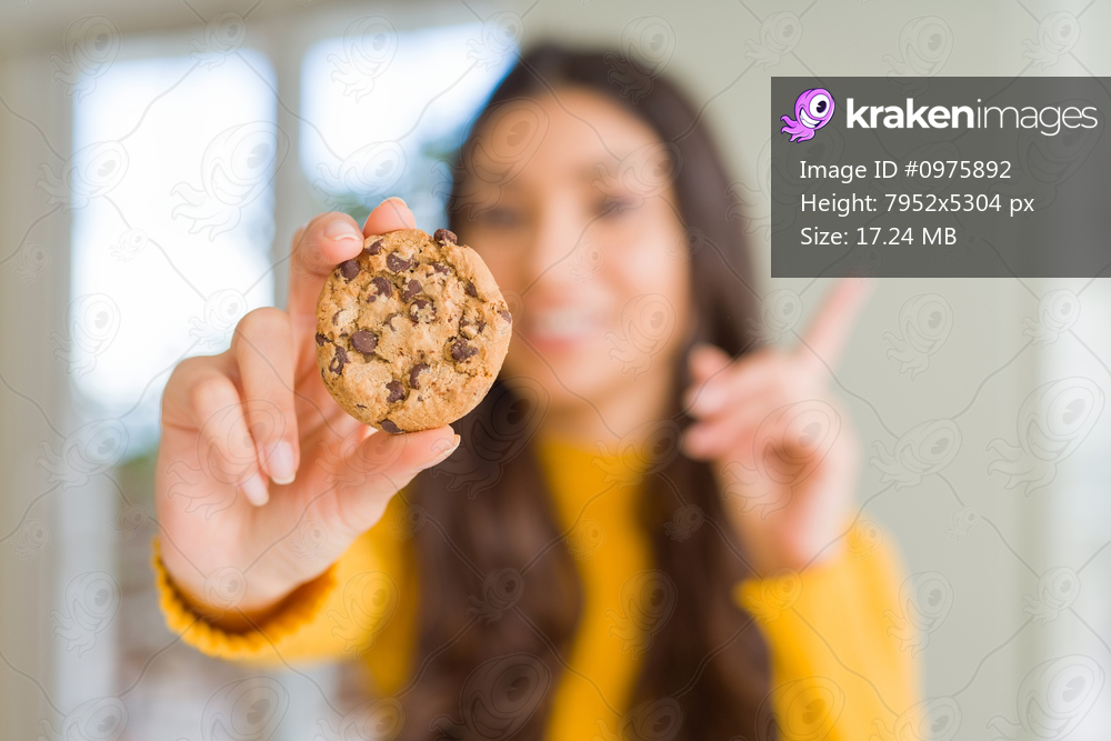 Young woman eating chocolate chips cookies at home surprised with an idea or question pointing finger with happy face, number one