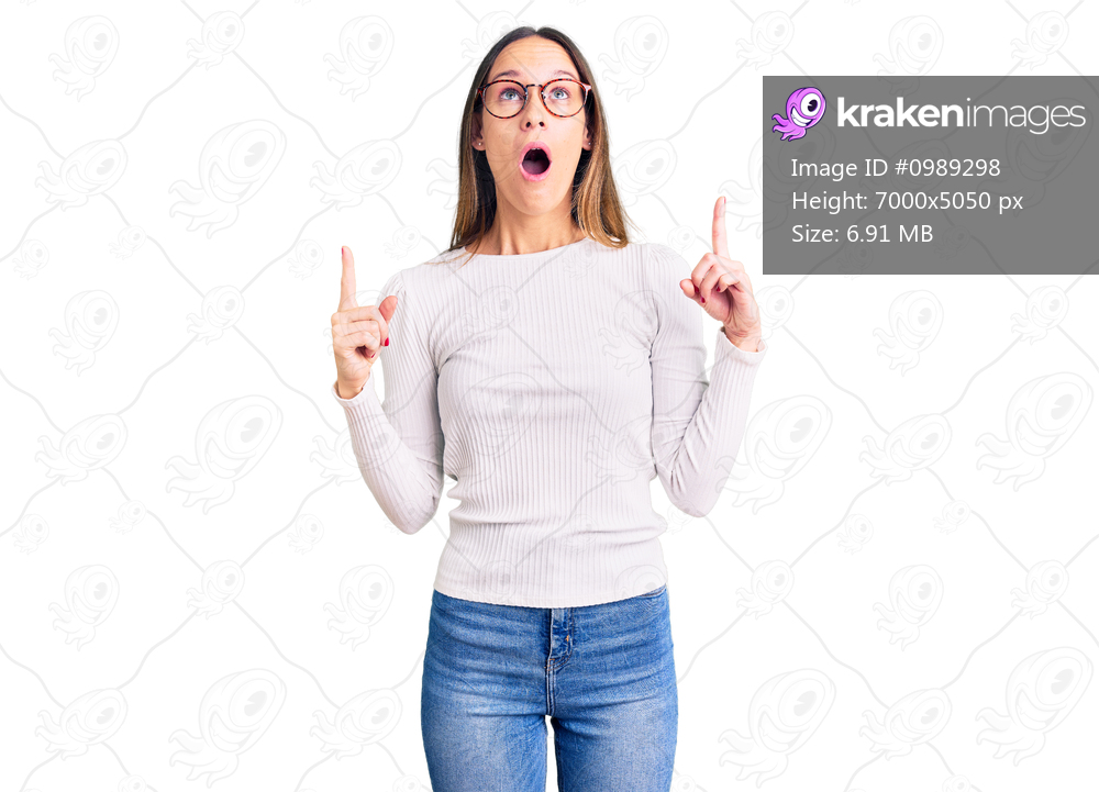 Beautiful brunette young woman wearing casual white sweater and glasses amazed and surprised looking up and pointing with fingers and raised arms. 