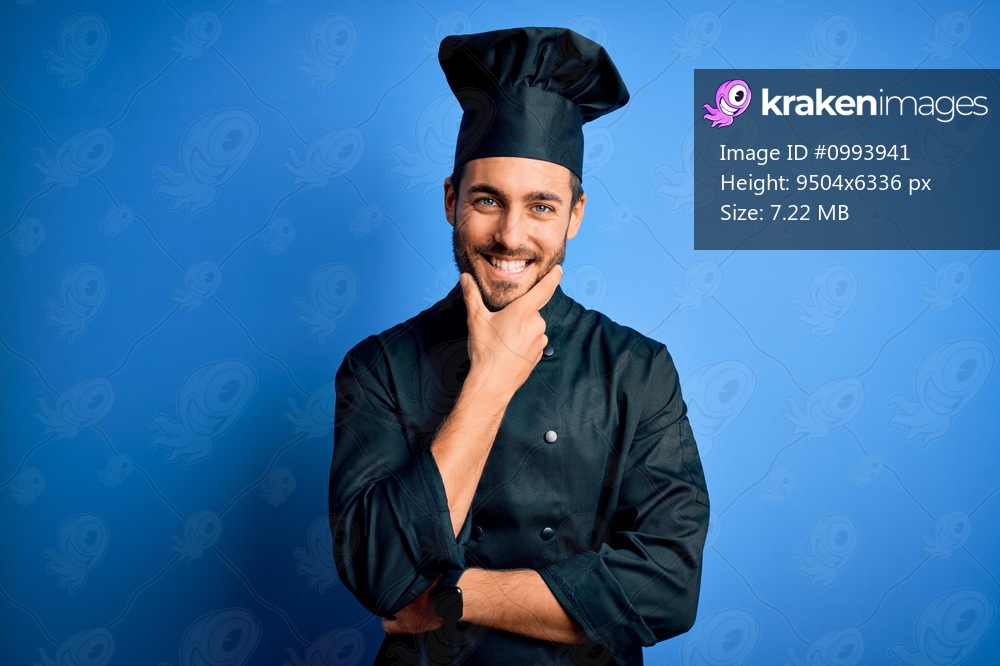 Young handsome chef man with beard wearing cooker uniform and hat over blue background looking confident at the camera smiling with crossed arms and hand raised on chin. Thinking positive.