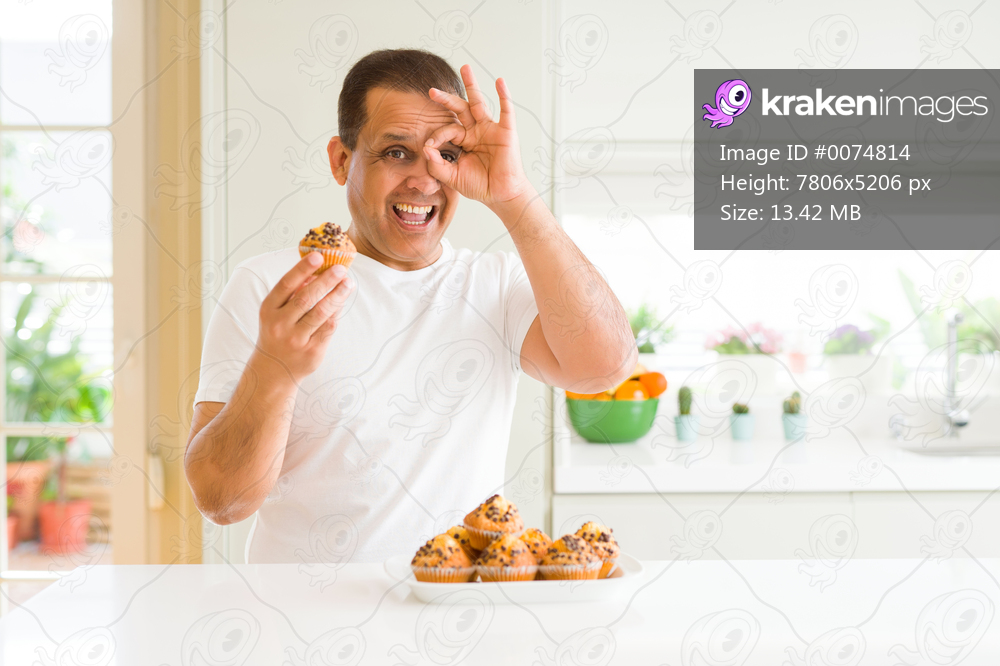 Middle age man eating chocolate chip muffins at home with happy face smiling doing ok sign with hand on eye looking through fingers