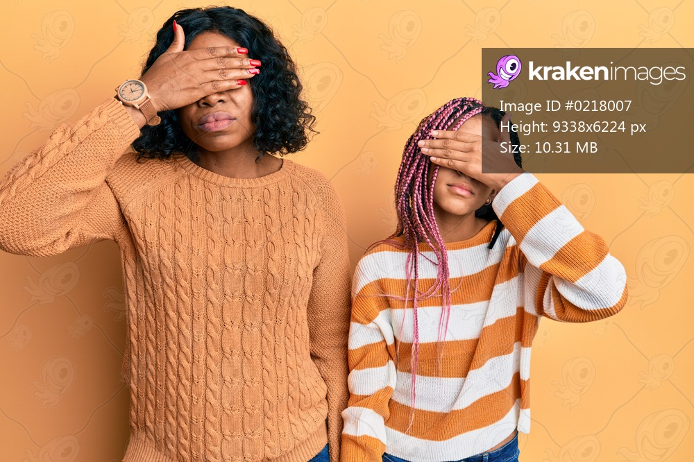 Beautiful african american mother and daughter wearing wool winter sweater covering eyes with hand, looking serious and sad. sightless, hiding and rejection concept 