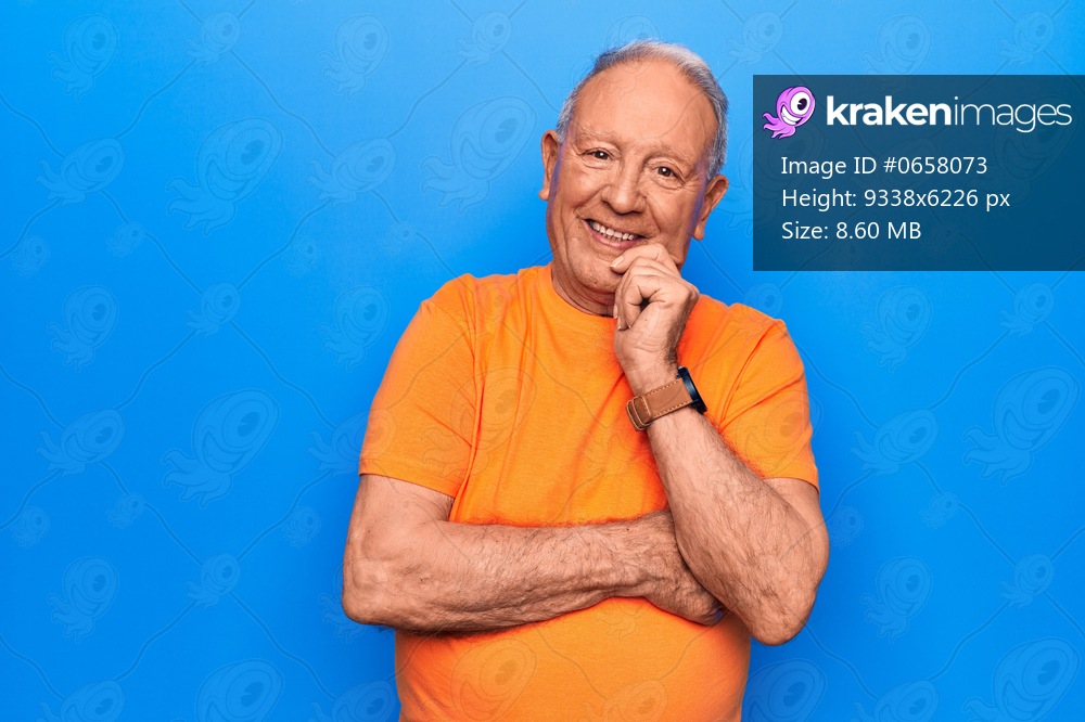 Senior handsome grey-haired man wearing casual t-shirt standing over blue background smiling looking confident at the camera with crossed arms and hand on chin. Thinking positive.