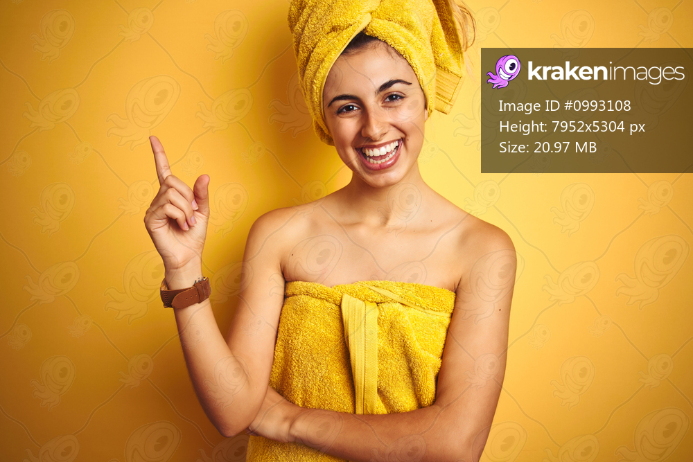 Young beautiful woman wearing a shower towel after bath over yellow isolated background with a big smile on face, pointing with hand and finger to the side looking at the camera.
