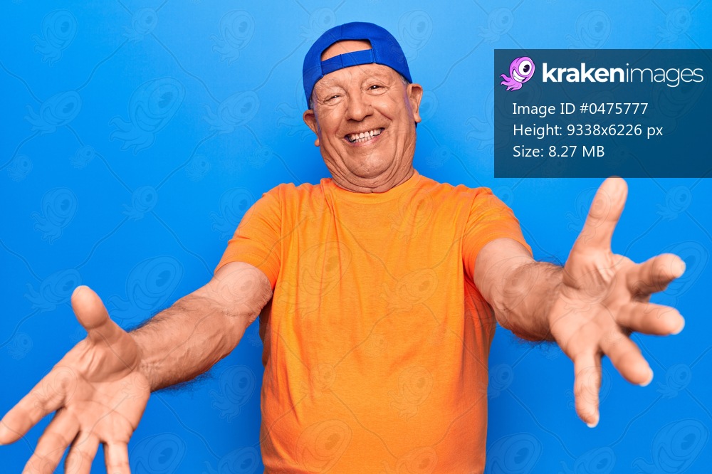 Senior handsome grey-haired man wearing casual t-shirt and cap over blue background looking at the camera smiling with open arms for hug. Cheerful expression embracing happiness.
