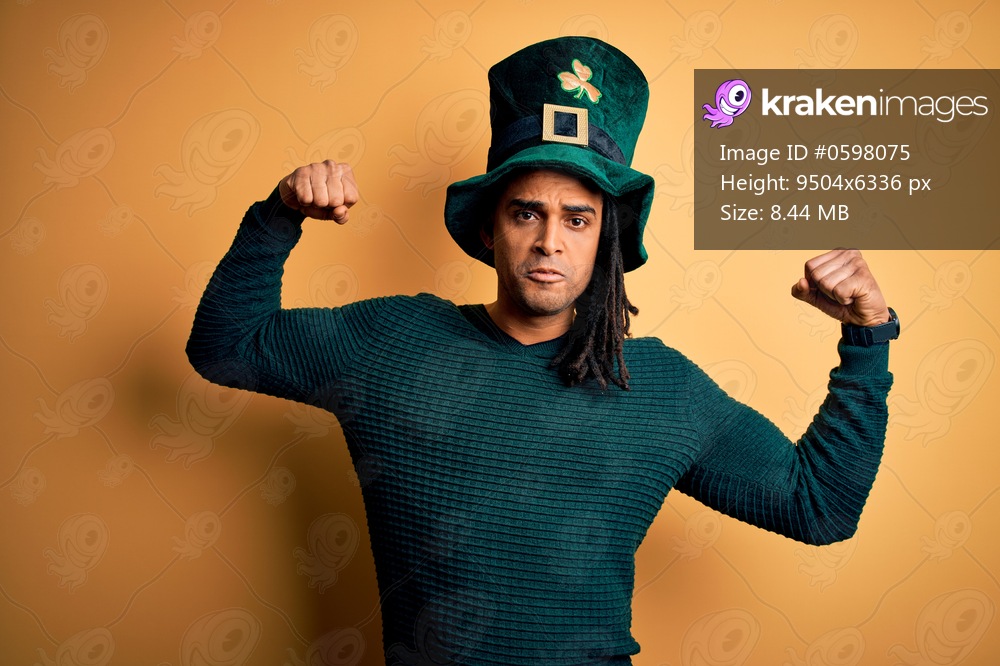 Young african american man wearing green hat with clover celebrating saint patricks day showing arms muscles smiling proud. Fitness concept.