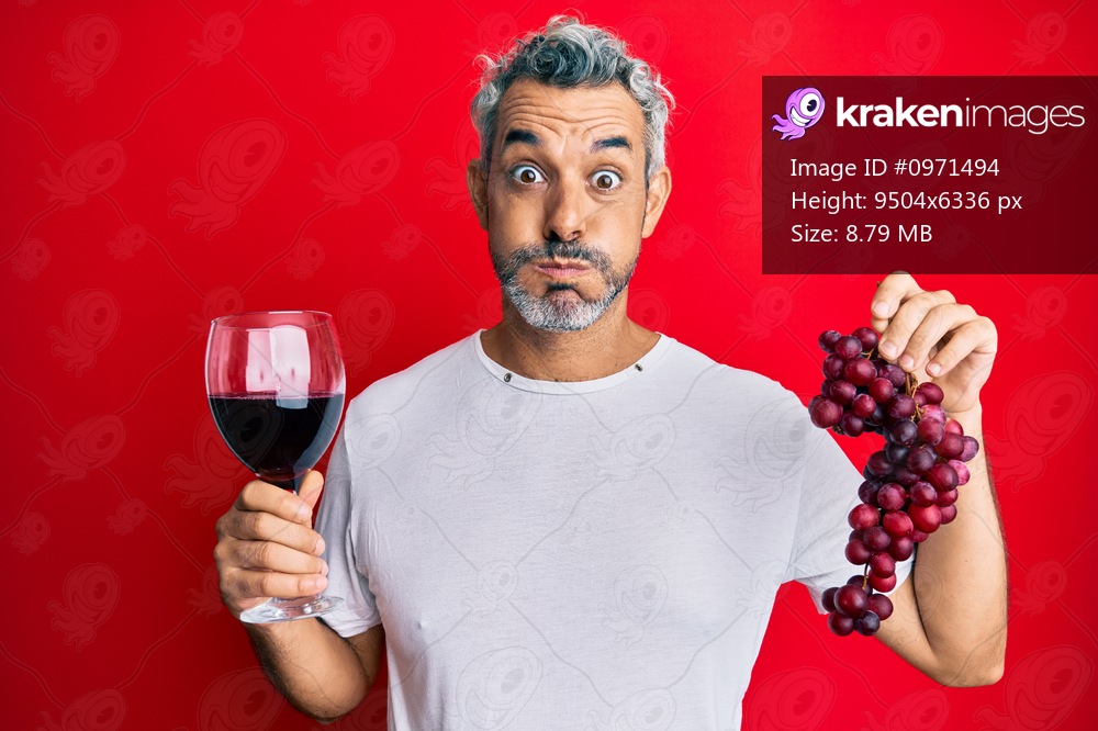 Middle age grey-haired man holding branch of fresh grapes and red wine puffing cheeks with funny face. mouth inflated with air, catching air. 