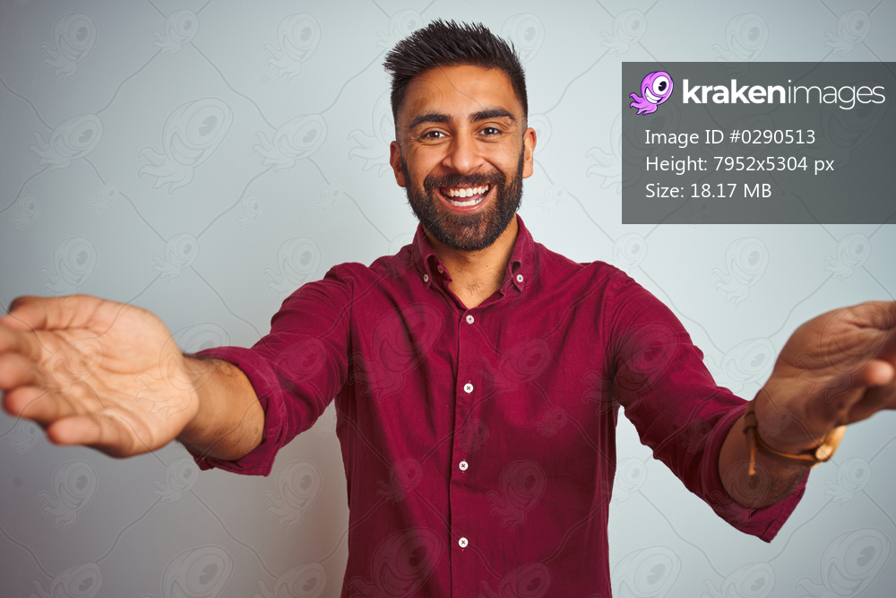 Young indian man wearing red elegant shirt standing over isolated grey background smiling cheerful offering hands giving assistance and acceptance.