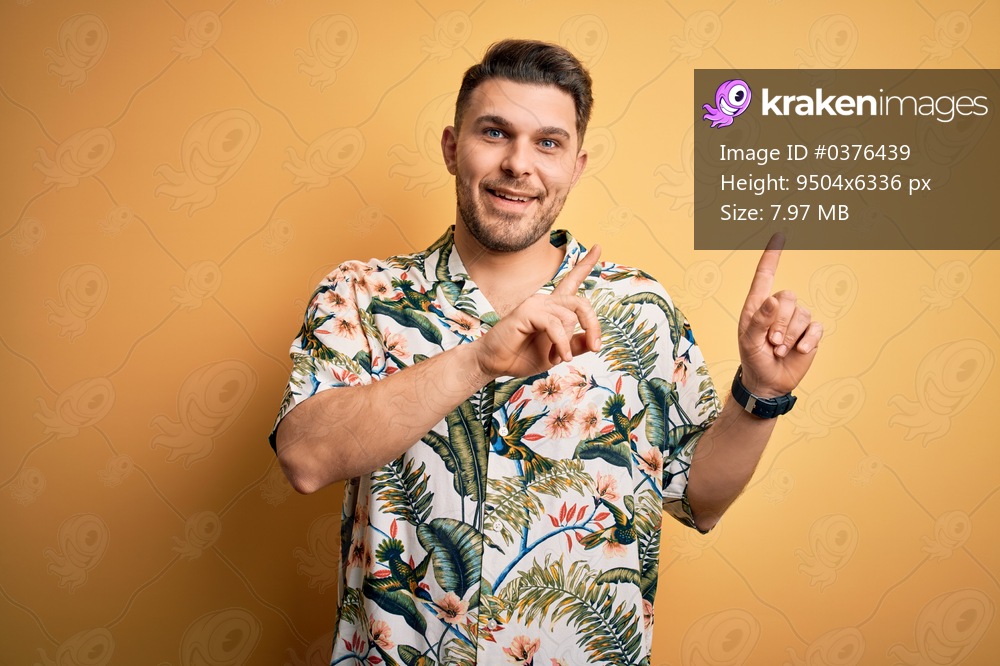 Young man with blue eyes on vacation wearing floral summer shirt over yellow background smiling and looking at the camera pointing with two hands and fingers to the side.