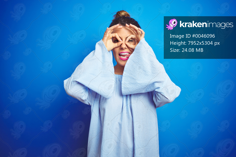 Young beautiful woman wearing bun hairstyle over blue isolated background doing ok gesture like binoculars sticking tongue out, eyes looking through fingers. Crazy expression.