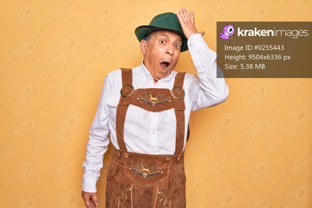 Senior grey-haired man wearing german traditional octoberfest suit over yellow background surprised with hand on head for mistake, remember error. Forgot, bad memory concept.