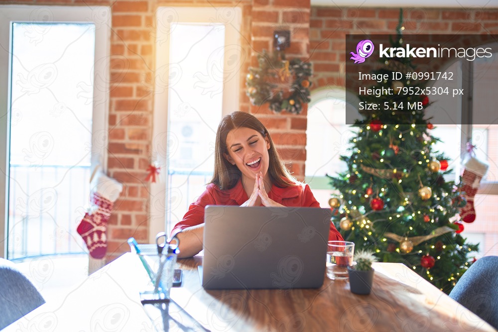Beautiful woman sitting at the table working with laptop at home around christmas tree Hands together and fingers crossed smiling relaxed and cheerful. Success and optimistic