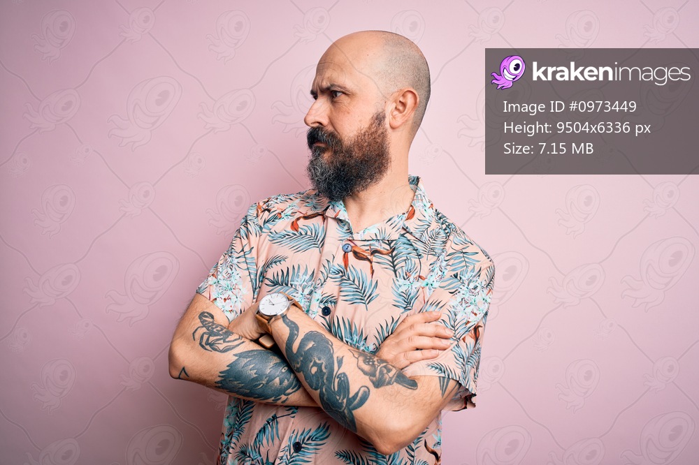 Handsome bald man with beard and tattoo wearing casual floral shirt over pink background looking to the side with arms crossed convinced and confident