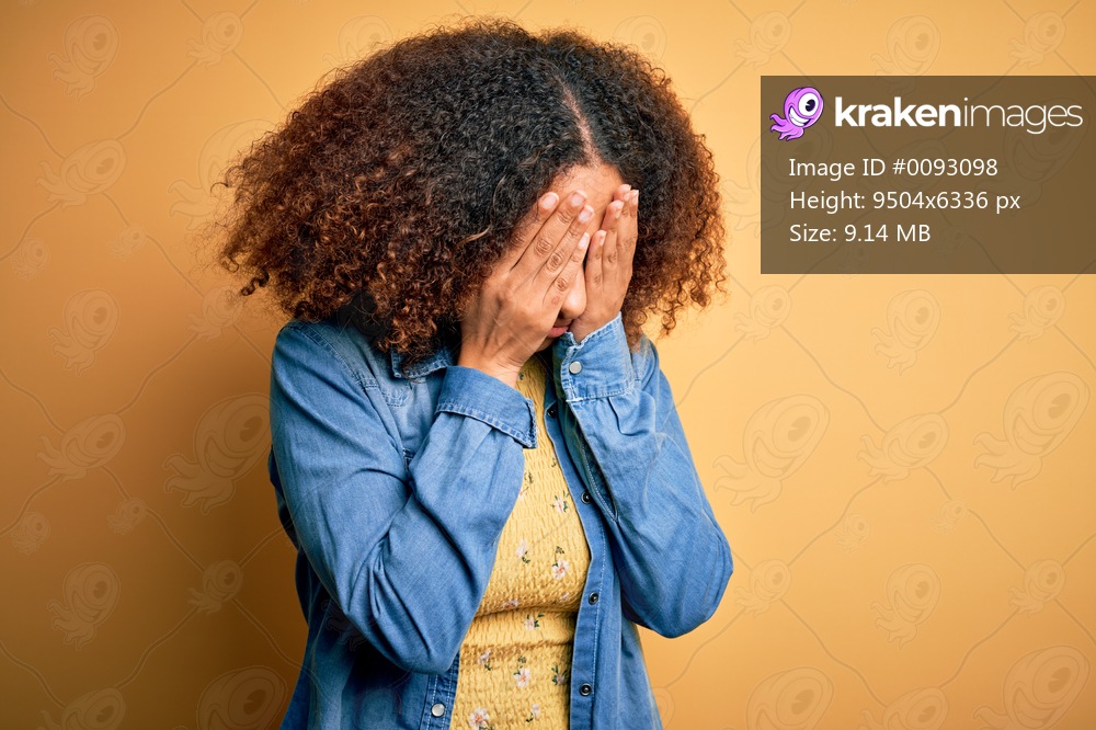 Young african american woman with afro hair wearing casual denim shirt over yellow background with sad expression covering face with hands while crying. Depression concept.