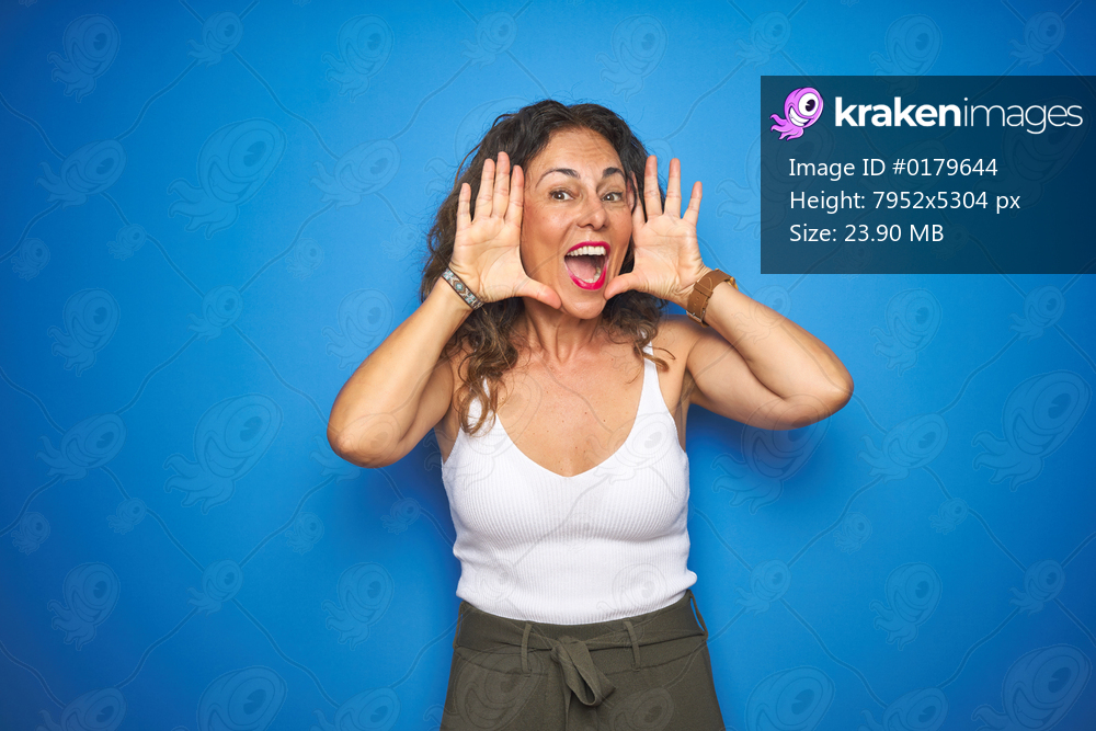 Middle age senior woman with curly hair standing over blue isolated background Smiling cheerful playing peek a boo with hands showing face. Surprised and exited