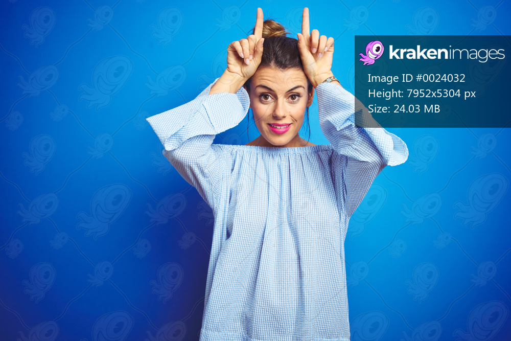 Young beautiful woman wearing bun hairstyle over blue isolated background doing funny gesture with finger over head as bull horns