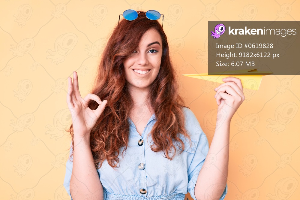 Young beautiful woman holding paper airplane doing ok sign with fingers, smiling friendly gesturing excellent symbol 