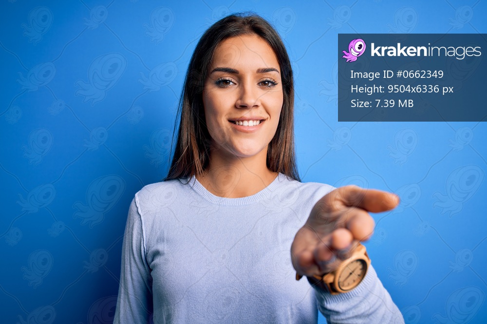 Young beautiful brunette woman wearing casual sweater standing over blue background smiling cheerful offering palm hand giving assistance and acceptance.