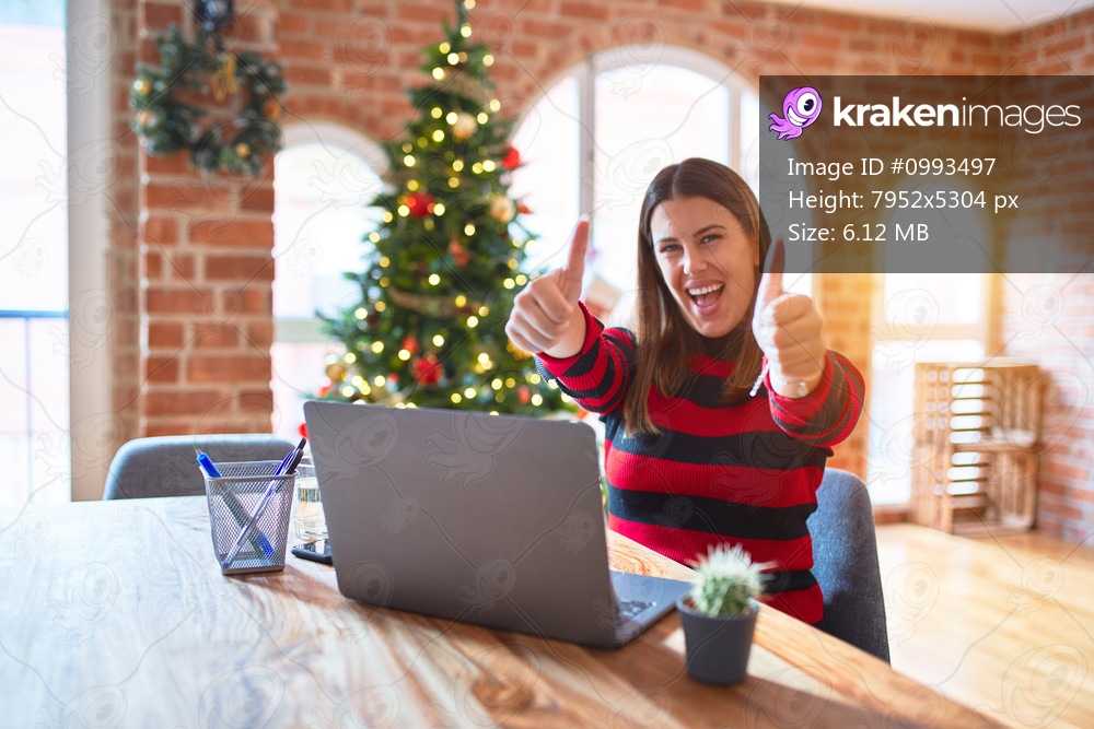 Beautiful woman sitting at the table working with laptop at home around christmas tree approving doing positive gesture with hand, thumbs up smiling and happy for success. Winner gesture.