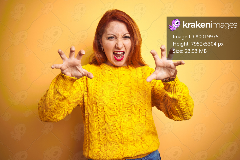 Beautiful redhead woman wearing winter sweater standing over isolated yellow background smiling funny doing claw gesture as cat, aggressive and sexy expression