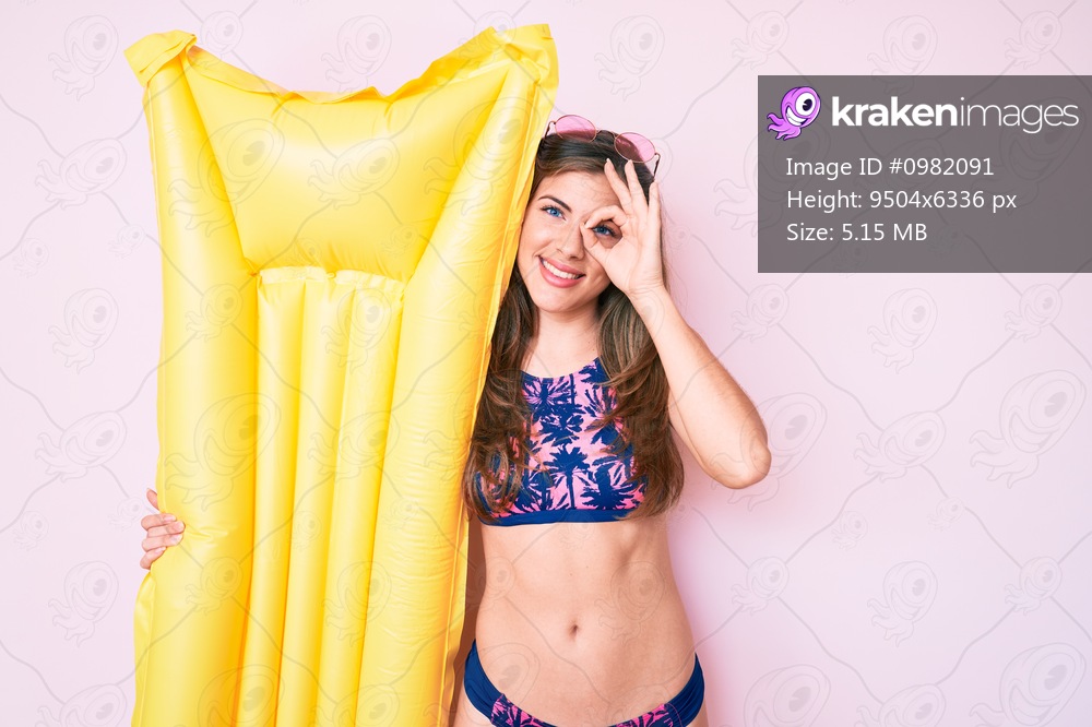 Beautiful young caucasian woman wearing bikini and holding summer matress float smiling happy doing ok sign with hand on eye looking through fingers 