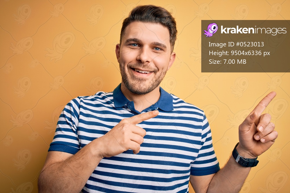 Young man with blue eyes wearing casual striped t-shirt over yellow background smiling and looking at the camera pointing with two hands and fingers to the side.