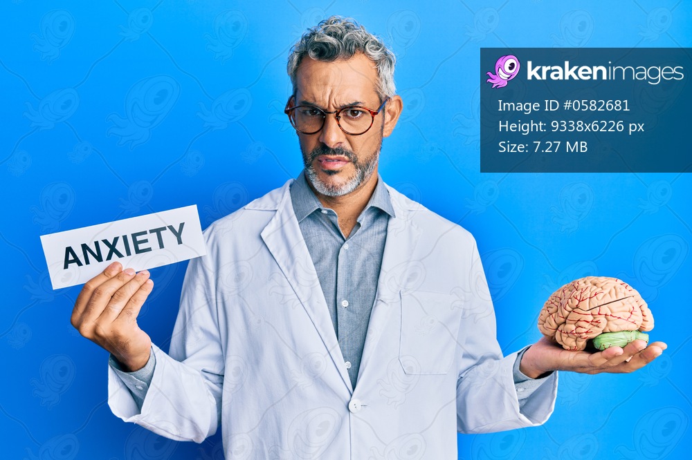 Middle age grey-haired man wearing doctor coat holding brain and anxiety message clueless and confused expression. doubt concept. 