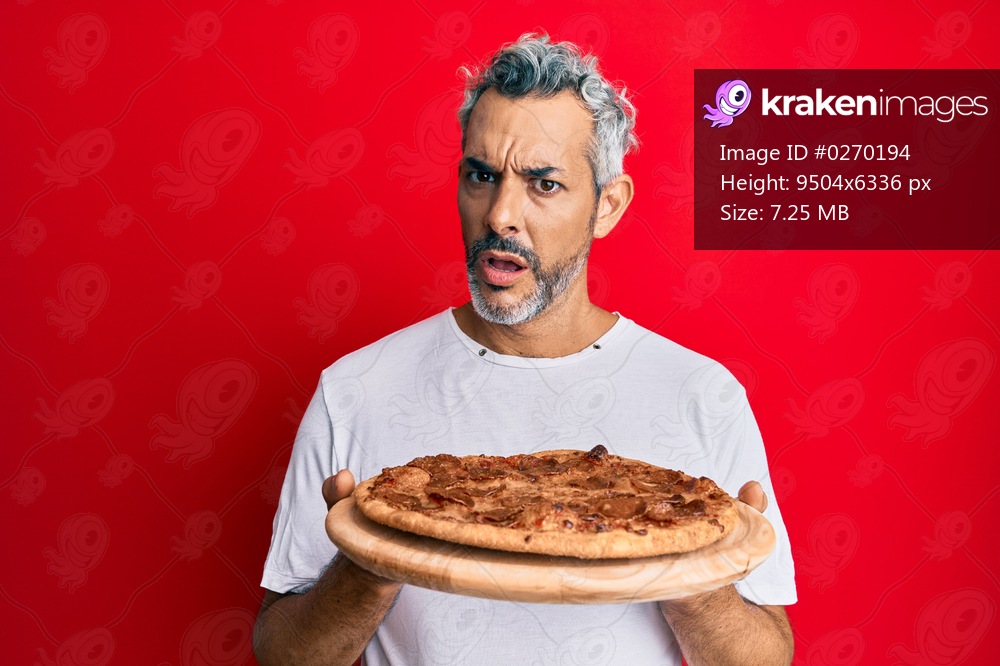 Middle age grey-haired man holding italian pizza in shock face, looking skeptical and sarcastic, surprised with open mouth 
