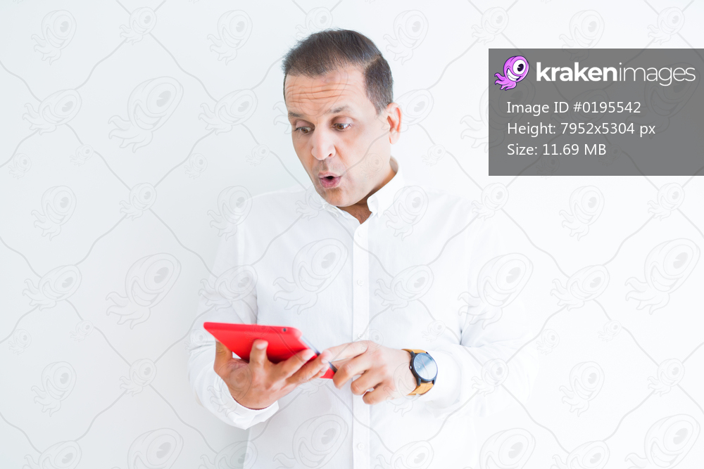 Middle age man using digital table over white wall scared in shock with a surprise face, afraid and excited with fear expression