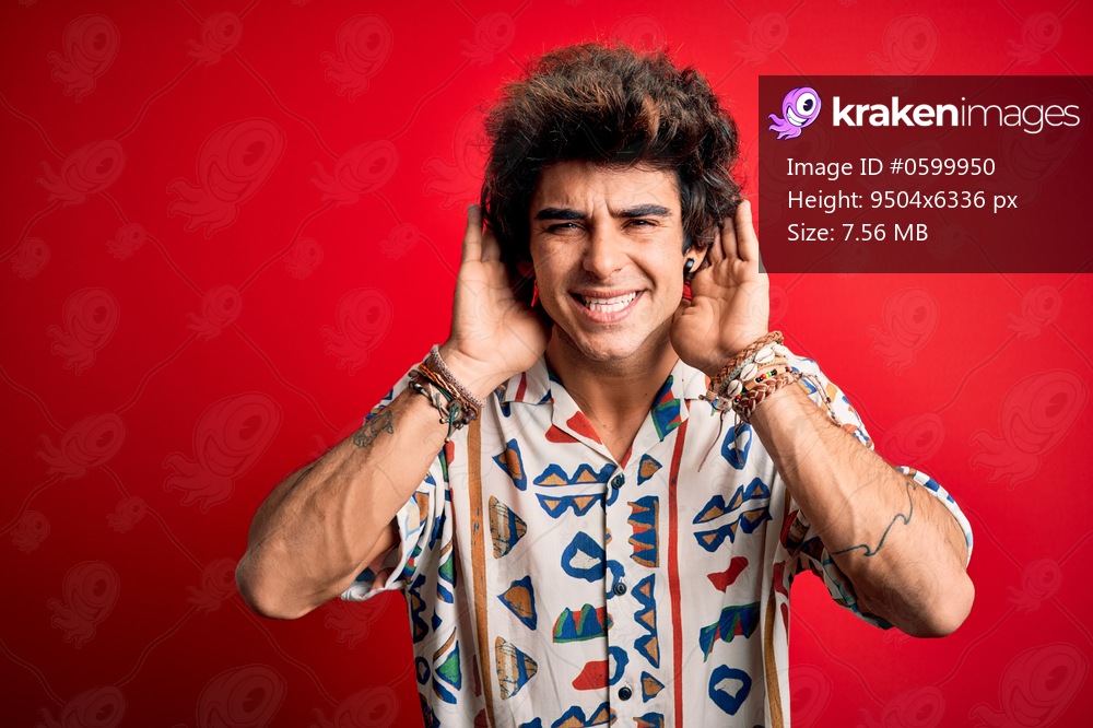 Young handsome man on vacation wearing summer shirt over isolated red background Trying to hear both hands on ear gesture, curious for gossip. Hearing problem, deaf