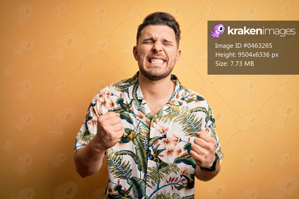 Young man with blue eyes on vacation wearing floral summer shirt over yellow background very happy and excited doing winner gesture with arms raised, smiling and screaming for success. Celebration concept.