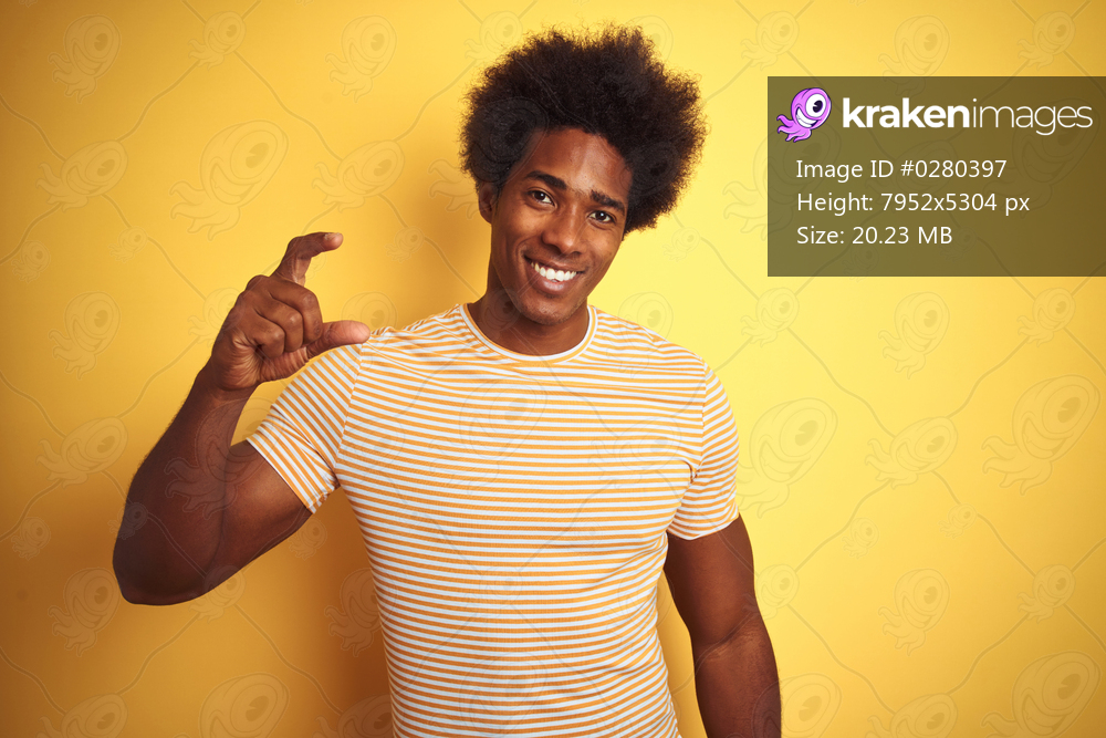 American man with afro hair wearing striped t-shirt standing over isolated yellow background smiling and confident gesturing with hand doing small size sign with fingers looking and the camera. Measure concept.