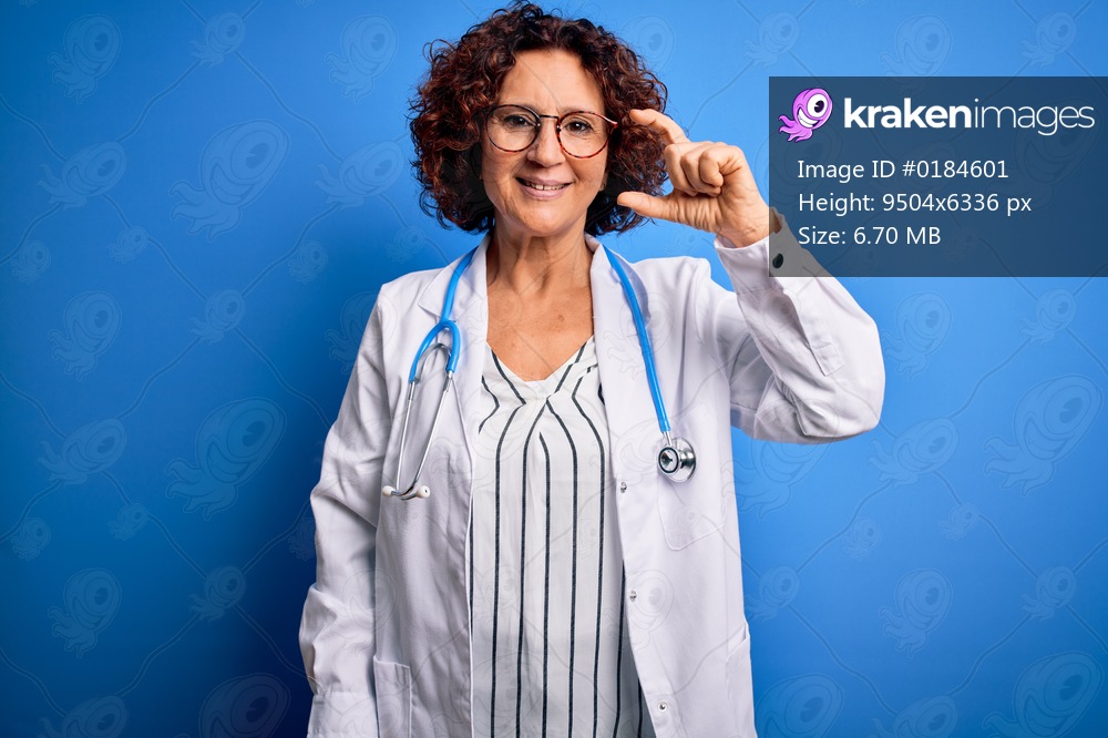 Middle age curly hair doctor woman wearing coat and stethoscope over blue background smiling and confident gesturing with hand doing small size sign with fingers looking and the camera. Measure concept.