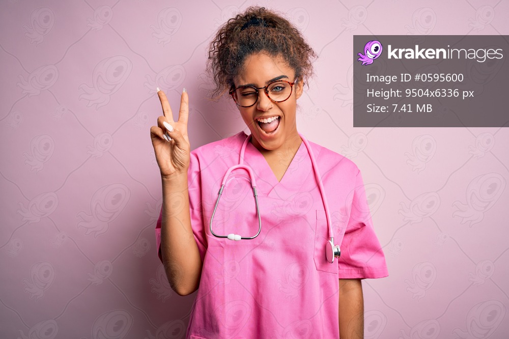 African american nurse girl wearing medical uniform and stethoscope over pink background smiling with happy face winking at the camera doing victory sign. Number two.