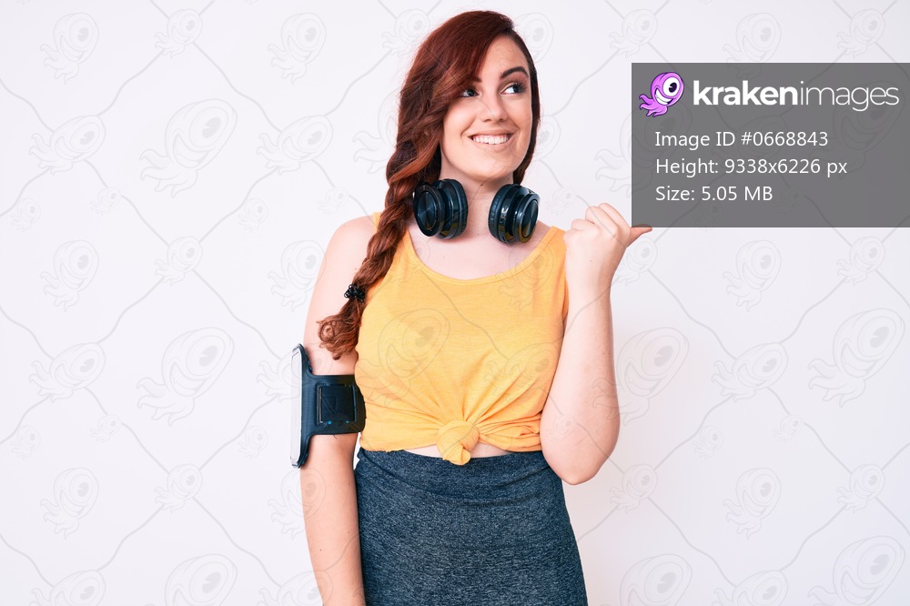 Young beautiful woman wearing gym clothes and using headphones smiling with happy face looking and pointing to the side with thumb up. 