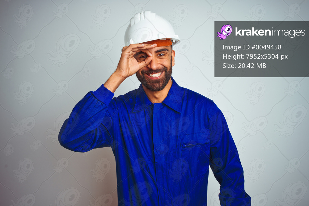 Handsome indian worker man wearing uniform and helmet over isolated white background doing ok gesture with hand smiling, eye looking through fingers with happy face.