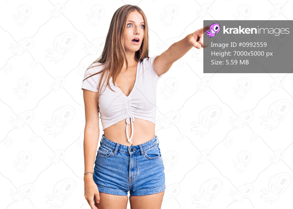 Young beautiful blonde woman wearing casual white tshirt pointing with finger surprised ahead, open mouth amazed expression, something on the front 