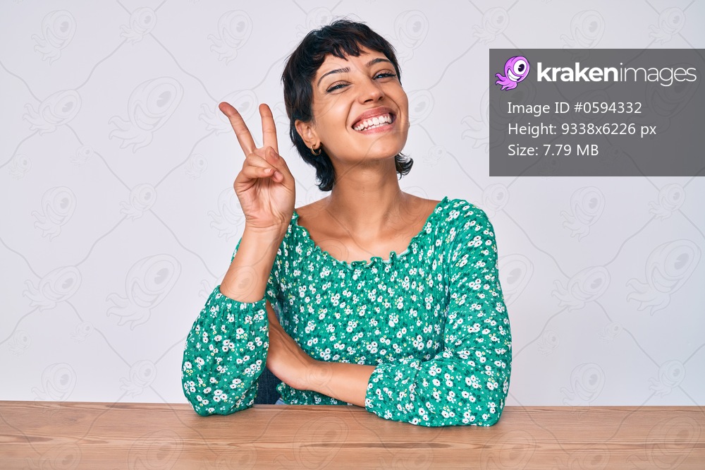 Beautiful brunettte woman wearing casual clothes sitting on the table smiling with happy face winking at the camera doing victory sign. number two. 