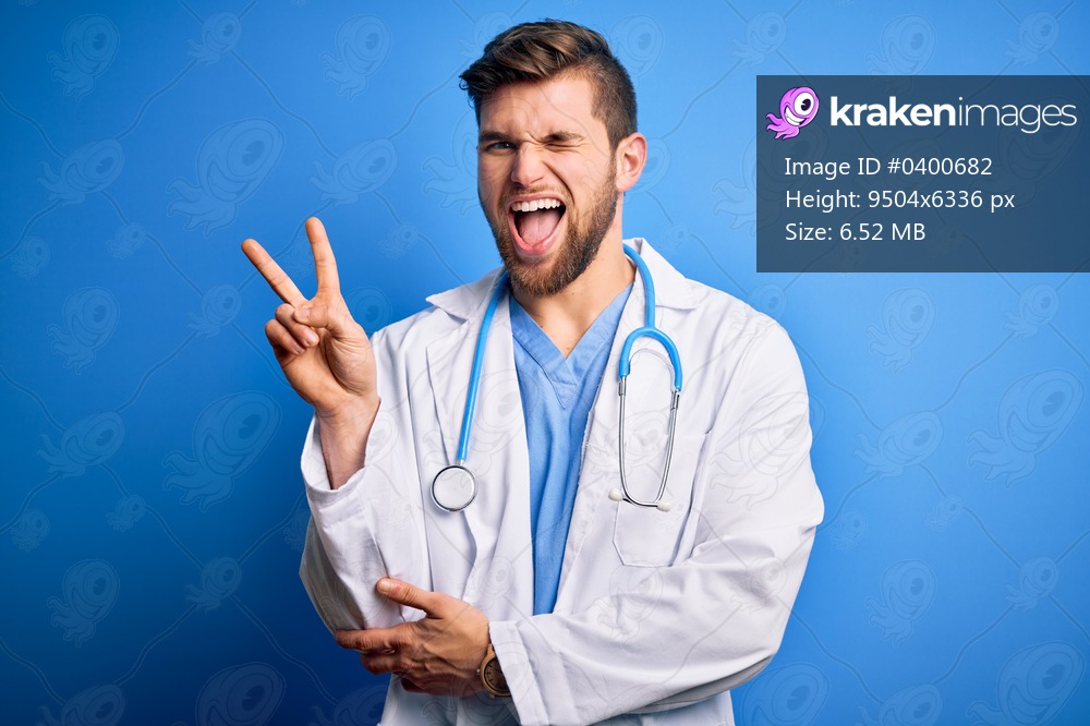 Young blond doctor man with beard and blue eyes wearing white coat and stethoscope smiling with happy face winking at the camera doing victory sign. Number two.