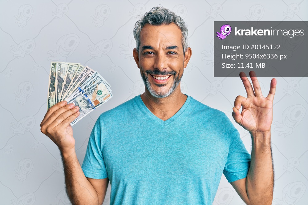 Middle age grey-haired man holding dollars doing ok sign with fingers, smiling friendly gesturing excellent symbol 