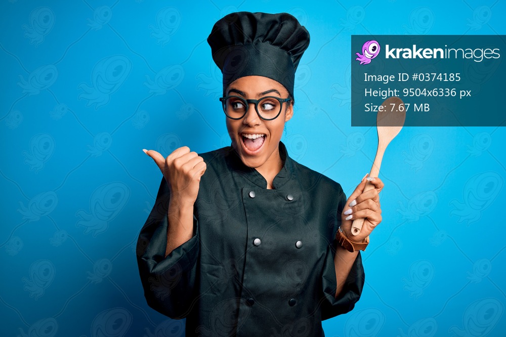Young african american chef girl wearing cooker uniform and hat holding wooden spoon tool pointing and showing with thumb up to the side with happy face smiling