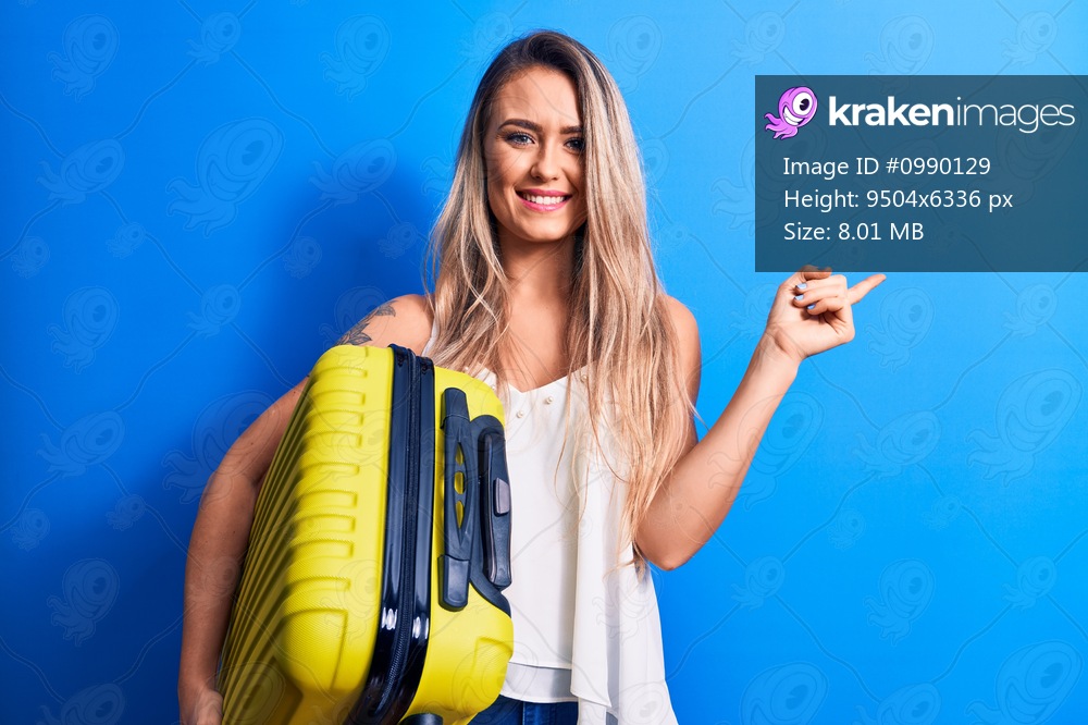 Young beautiful blonde tourist woman on vacation holding suitcase over blue background smiling happy pointing with hand and finger to the side