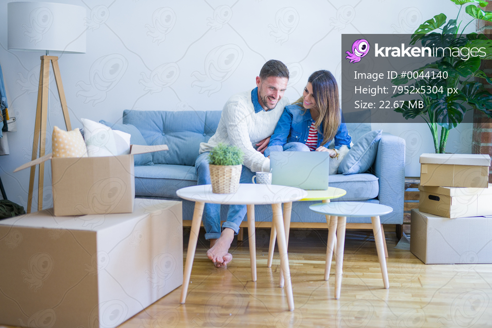 Young beautiful couple sitting on the sofa drinking cup of coffee using laptop at new home around cardboard boxes