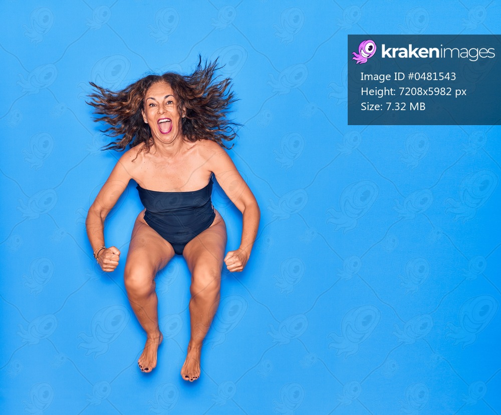 Middle age beautiful hispanic woman on vacation wearing swimsuit smiling happy. Jumping with smile on face celebrating over isolated blue background