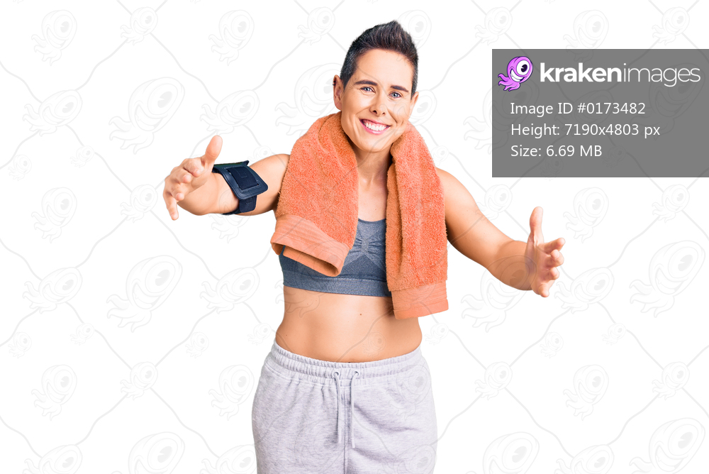 Young woman with short hair wearing sportswear and towel using smartphone looking at the camera smiling with open arms for hug. cheerful expression embracing happiness. 