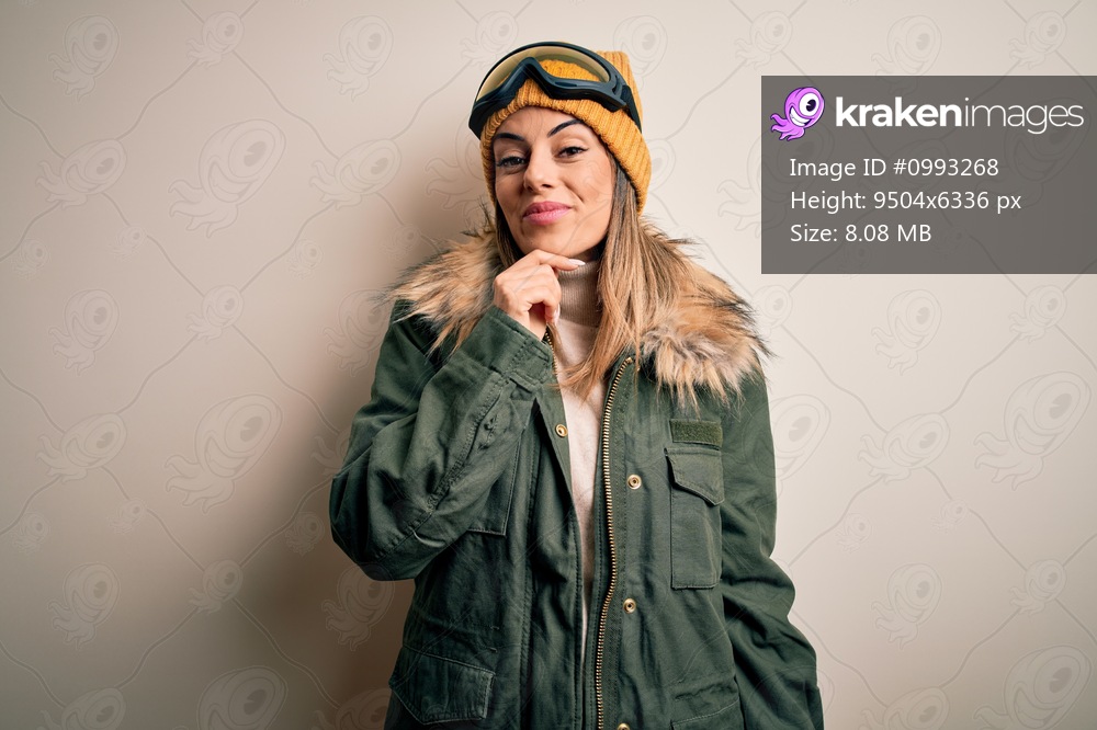 Young brunette skier woman wearing snow clothes and ski goggles over white background looking confident at the camera smiling with crossed arms and hand raised on chin. Thinking positive.